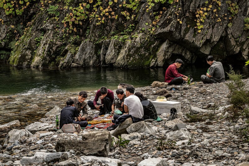 A group of people are sitting on rocky ground by a river, enjoying a picnic. They are surrounded by lush greenery and large rock formations. Food items and picnic supplies are spread out on blankets in front of them.