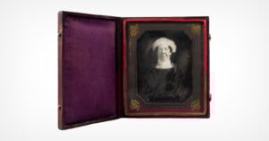 A framed, black and white photograph of an elderly woman wearing a headscarf, displayed in an antique case with a purple velvet lining and red trim. The case is open, showcasing the portrait.