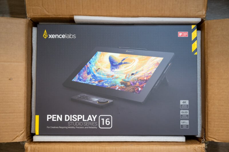 The image shows the packaging of a Xencelabs Pen Display Studio Series 16. The box features a vibrant illustration on the tablet screen, with text highlighting its 4K resolution, OLED display, and various features for creative professionals. The box is inside a cardboard shipping container.