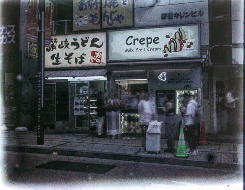 A grainy, vintage-looking photograph of a bustling city street scene with pedestrians walking past shops, notably featuring signs for crepes and milk soft cream in both english and japanese.