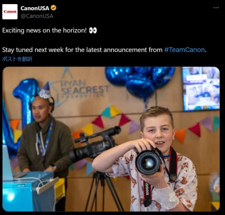 A young boy excitedly holds a camera, focusing intently in a room decorated with blue balloons and banners, while a man in the background looks on at an event booth marked "team canon.